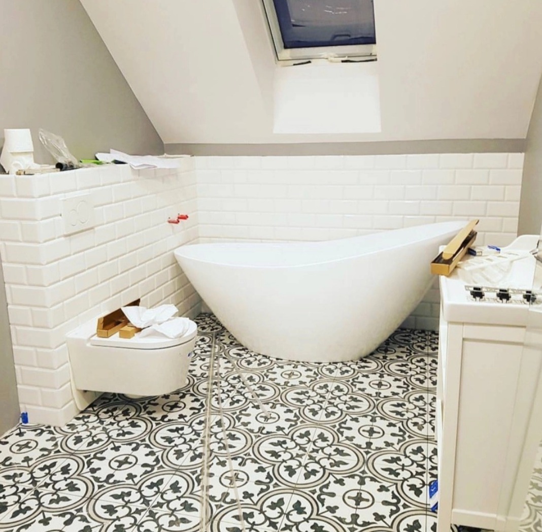 Nowoczesna łazienka na poddaszu w trakcie wykańczania: wolnostojąca wanna, podwieszana toaleta, białe płytki typu metro na ścianach, wzorzysta czarno-biała podłoga, okno dachowe.