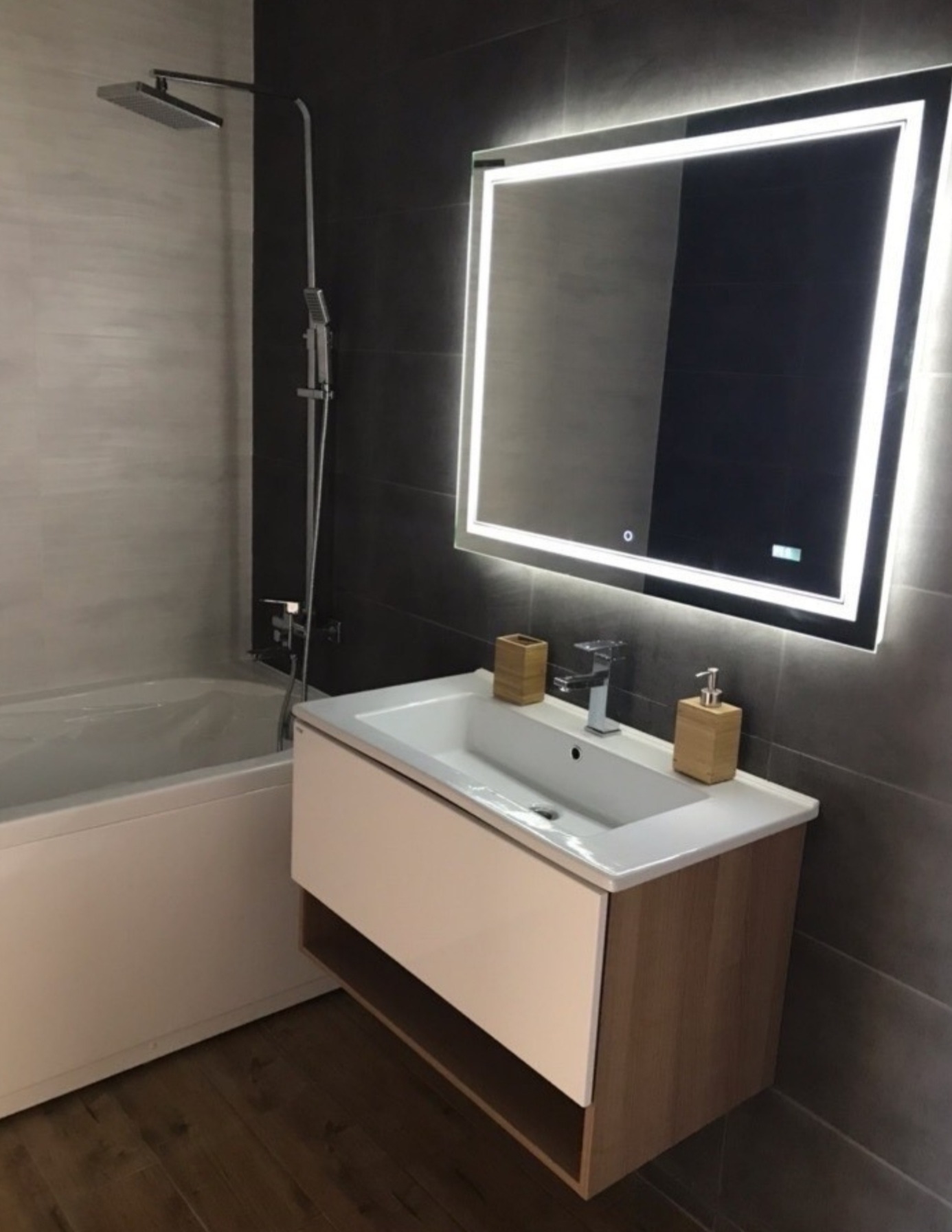 Nowoczesna łazienka z podświetlanym lustrem, białą umywalką z szafką z elementami drewna, szarą glazurą na ścianach oraz baterią umywalkową i dozownikiem mydła w kolorze srebrnym. W tle widoczna...