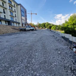 Przygotowanie podłoża z kruszywa pod drogę dojazdową do nowoczesnego budynku mieszkalnego, widoczny sprzęt budowlany i elementy betonowe obrzeża.