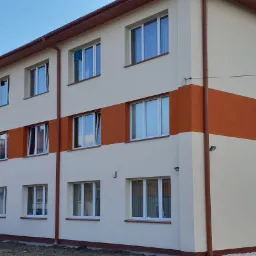 Odnowiony budynek wielorodzinny z beżową elewacją i poziomym pasem w kolorze terakoty, widoczne okna z białymi ramami i brązowe rynny, w tle komin i fragment placu budowy.