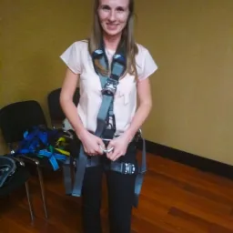 Uśmiechnięta kobieta w jasnej bluzce i czarnych spodniach, ubrana w szelki asekuracyjne z metalowymi klamrami, trzymająca karabińczyk, w tle krzesła i drewniana podłoga.