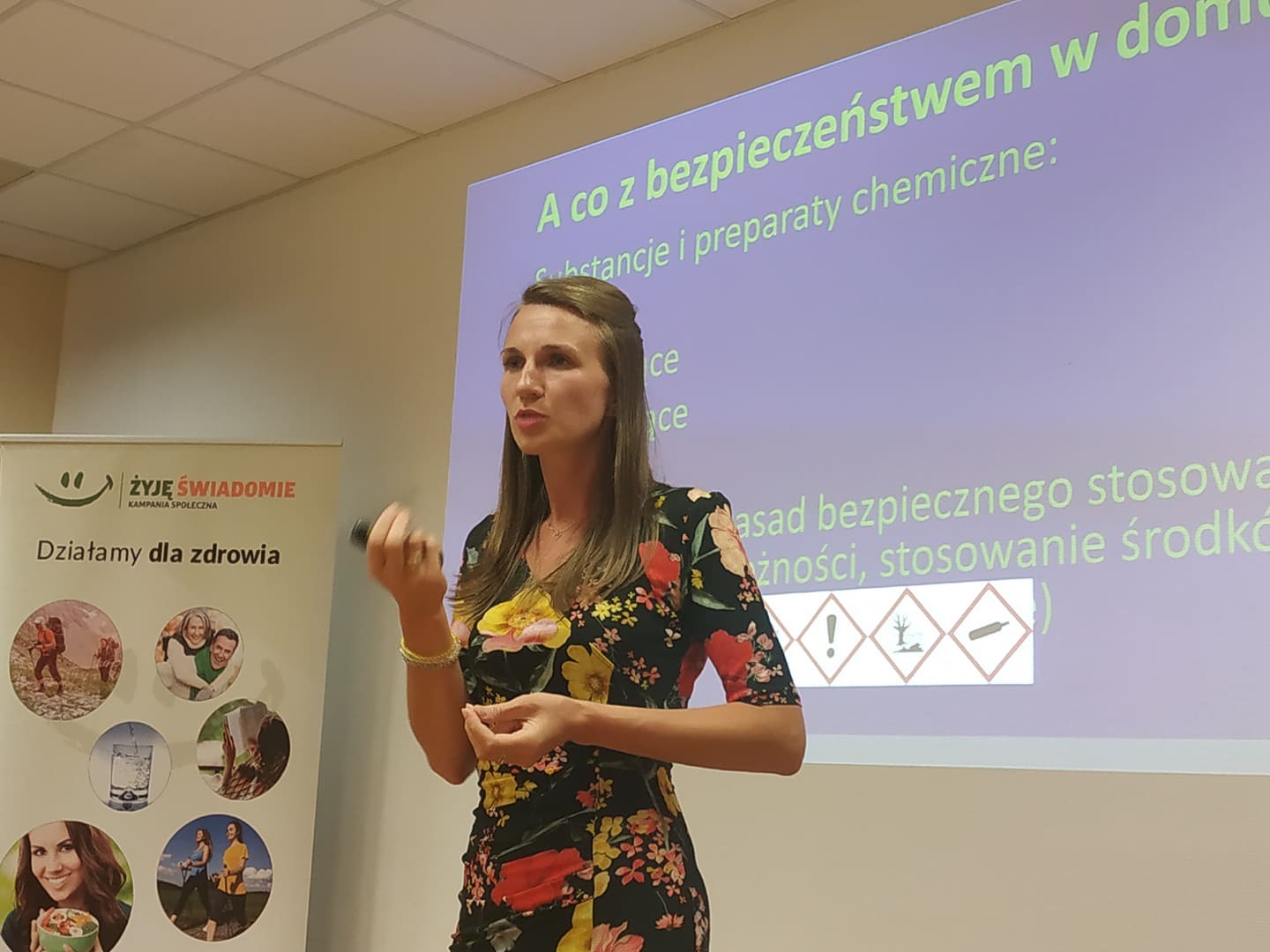 Kobieta w kwiecistej sukience prezentuje zasady bezpieczeństwa chemicznego podczas szkolenia, obok baner kampanii społecznej promującej zdrowy styl życia.