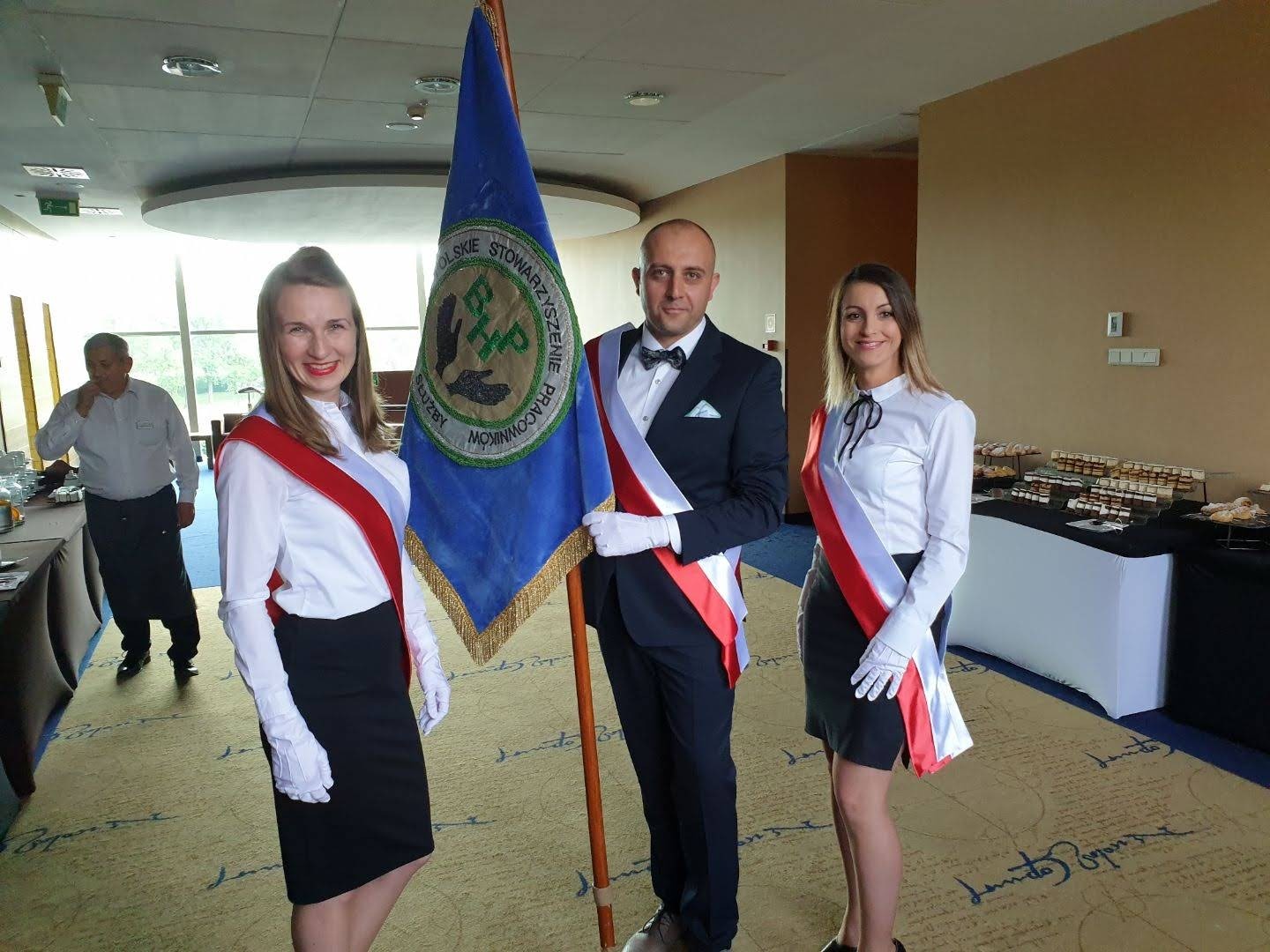 Trzy osoby w strojach galowych z szarfami w barwach narodowych stoją obok sztandaru na tle sali konferencyjnej z cateringiem.