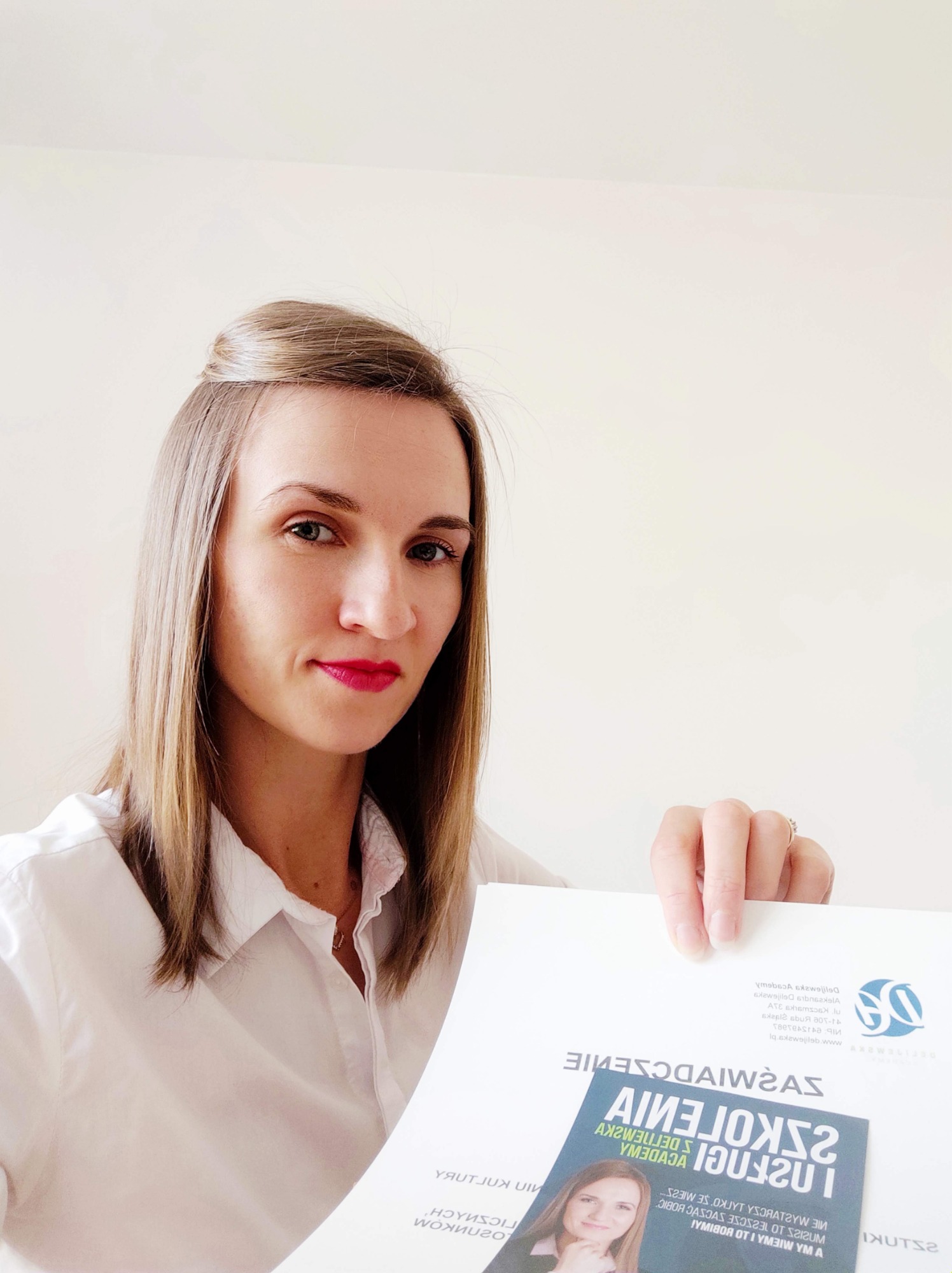 Kobieta w białej koszuli prezentuje certyfikat ukończenia szkolenia, z widocznym logo akademii i jej zdjęciem na ulotce.