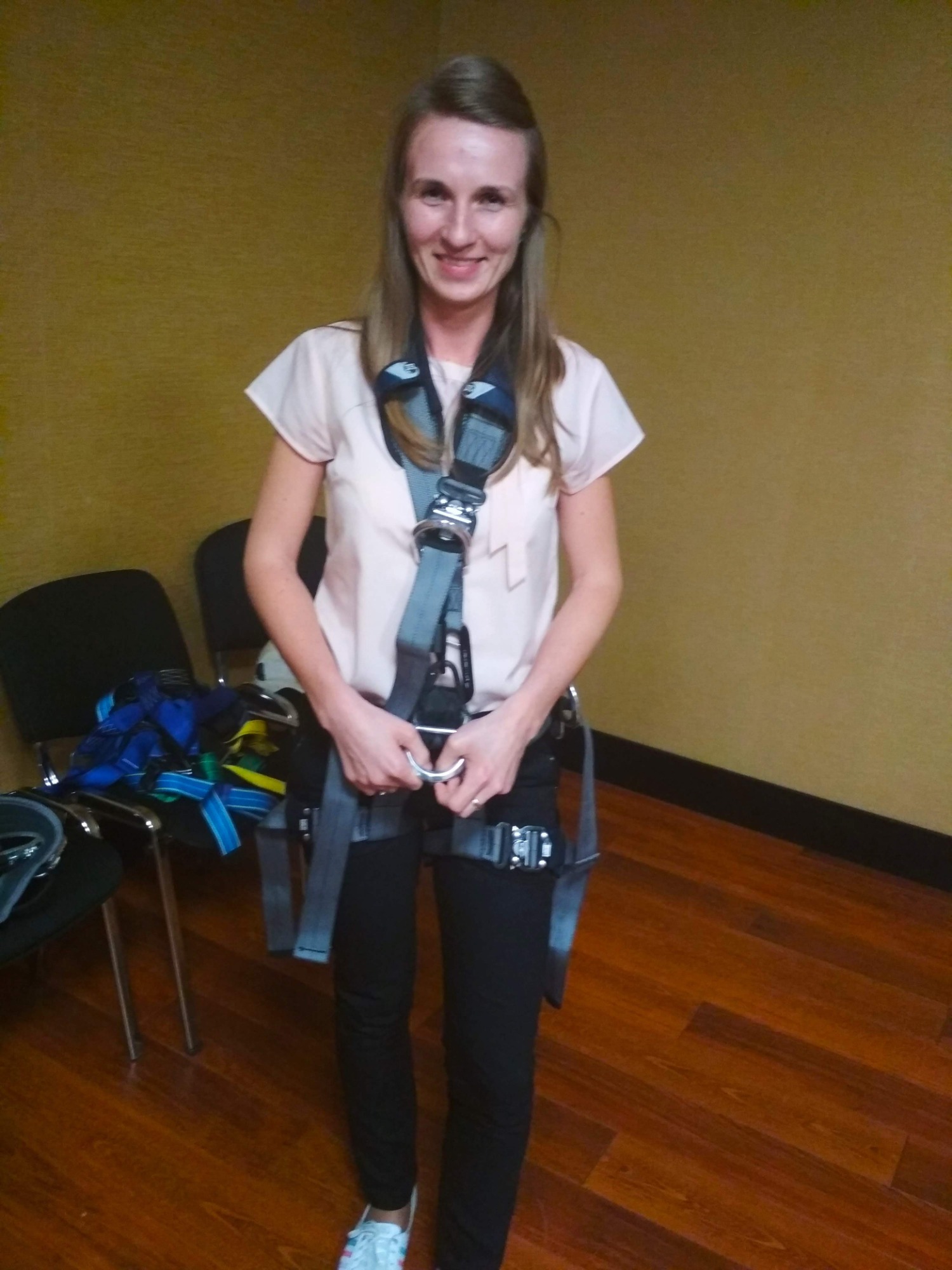 Uśmiechnięta kobieta w jasnej bluzce i czarnych spodniach, ubrana w szelki asekuracyjne z metalowymi klamrami, trzymająca karabińczyk, w tle krzesła i drewniana podłoga.