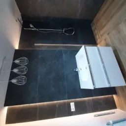 Nowoczesna łazienka z ciemnymi płytkami imitującymi marmur, drewnianą podłogą, minimalistyczną białą umywalką z szafką, designerską lampą wiszącą i chromowaną armaturą prysznicową.