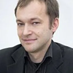 Portret mężczyzny w czarnej koszuli i marynarce, z delikatnym uśmiechem, na jasnym tle.