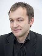 Portret mężczyzny w czarnej koszuli i marynarce, z delikatnym uśmiechem, na jasnym tle.