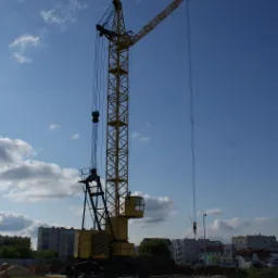 Żółty dźwig budowlany na tle błękitnego, lekko zachmurzonego nieba i zabudowań mieszkalnych. Widoczny fragment terenu budowy i inne maszyny budowlane.