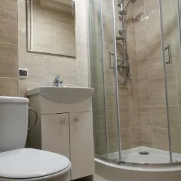 Jasna łazienka z kabiną prysznicową, umywalką z szafką i toaletą, wykończona płytkami w odcieniach beżu.