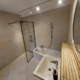 Nowoczesna łazienka po remoncie: kabina prysznicowa typu walk-in z czarną ramą, biała umywalka z drewnianym blatem, marmurowe płytki podłogowe i oświetlenie punktowe.