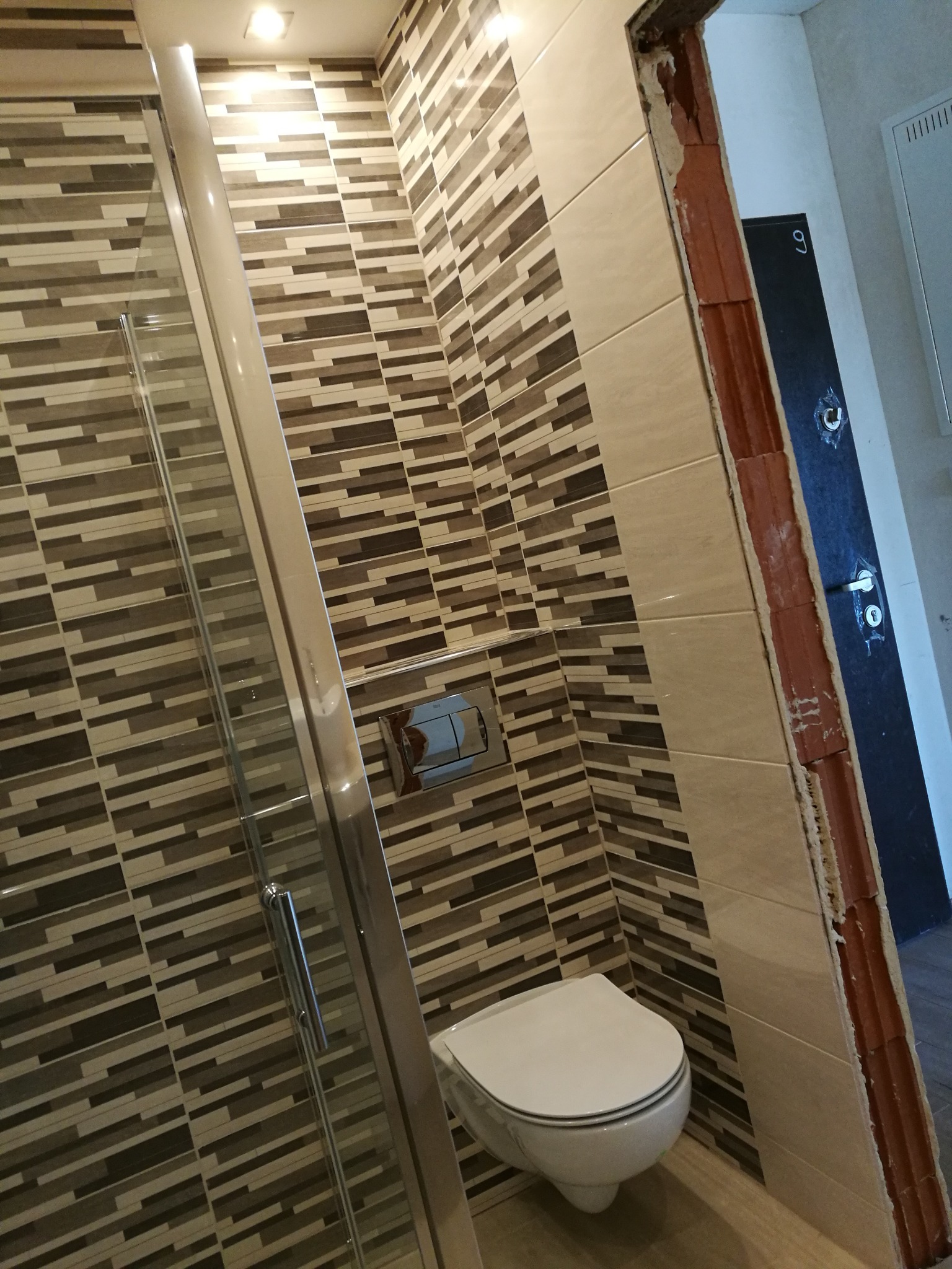 Ukończona łazienka w Pieńsku z kabiną prysznicową, podwieszaną toaletą i nowoczesnymi płytkami w odcieniach brązu i beżu, widoczna futryna drzwi z ceglanym obramowaniem.