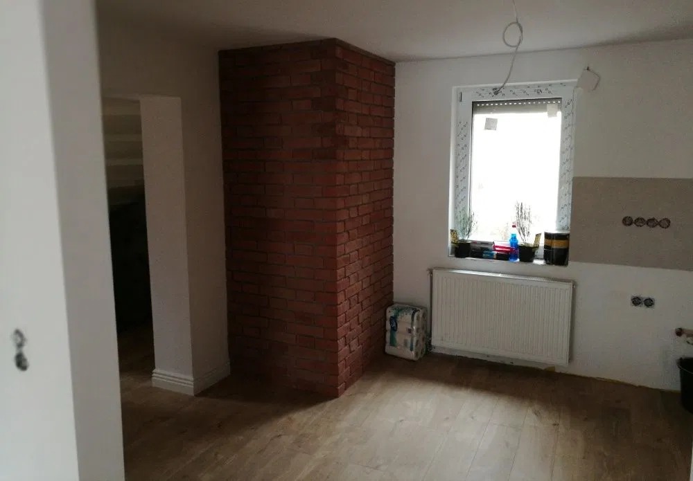 Fragment łazienki w trakcie remontu: ściana wykończona czerwoną cegłą, okno z grzejnikiem, częściowo położona podłoga z paneli, widoczne puszki elektryczne i niedokończone ściany.