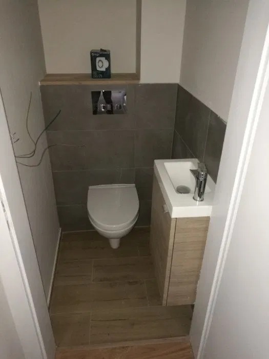 Mała łazienka po remoncie: podwieszana toaleta, umywalka z szafką w odcieniach drewna, szare płytki ścienne i drewnopodobna podłoga. Na półce nad toaletą pudełko z akcesoriami.