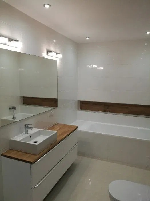 Jasna łazienka po remoncie: białe płytki, drewniane akcenty nad wanną i na blacie szafki umywalkowej, prostokątna umywalka nablatowa, duże lustro z oświetleniem.