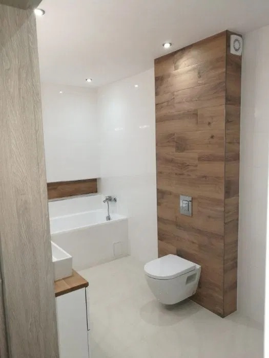 Nowoczesna łazienka z białą ceramiką, drewnopodobną ścianą z ukrytym systemem spłukiwania, wanną z zabudową i umywalką na drewnianym blacie.