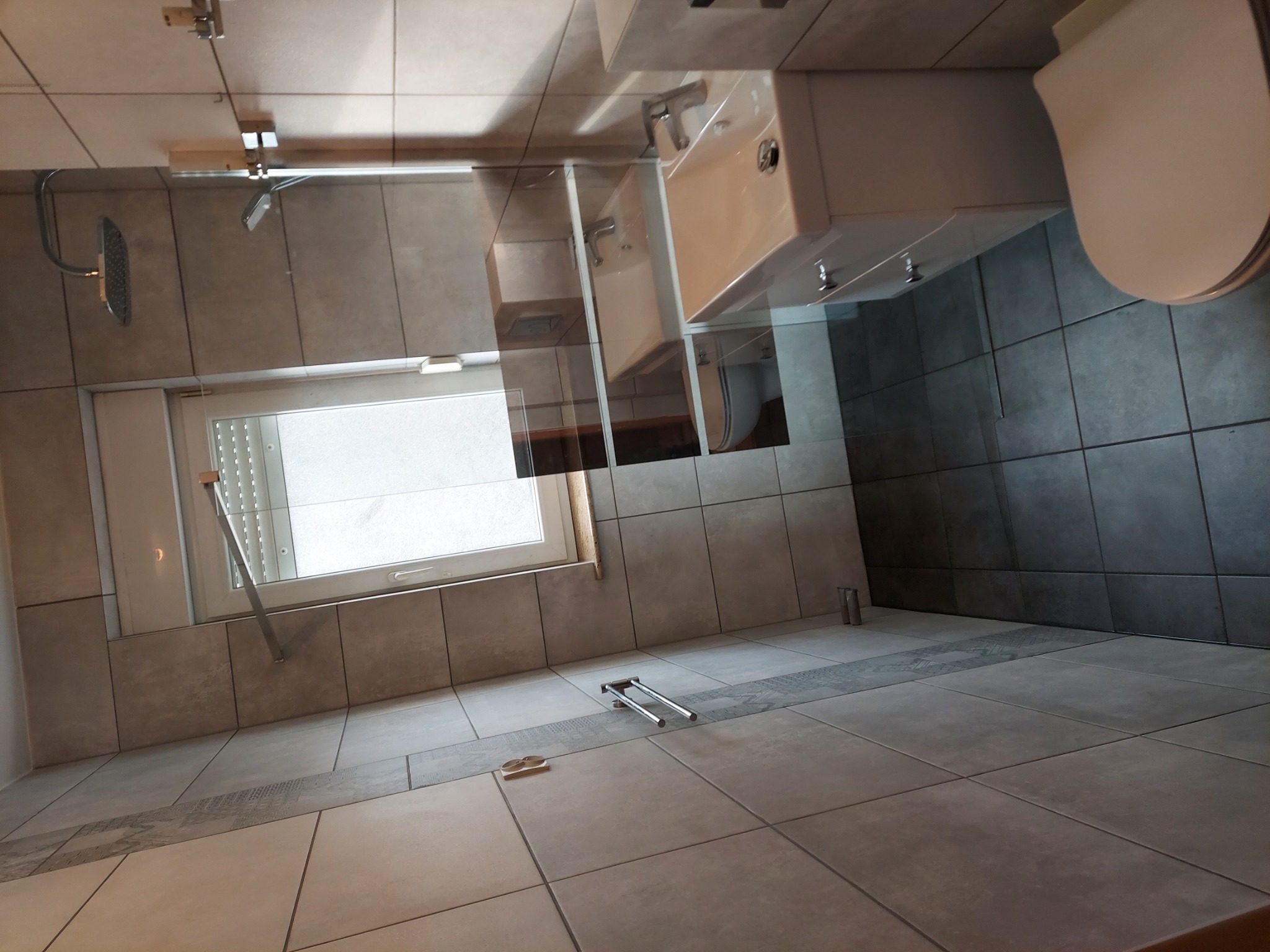 Wykończona łazienka z szarymi płytkami, wiszącą białą szafką z umywalką i toaletą, oświetlona naturalnym światłem z okna z roletami.