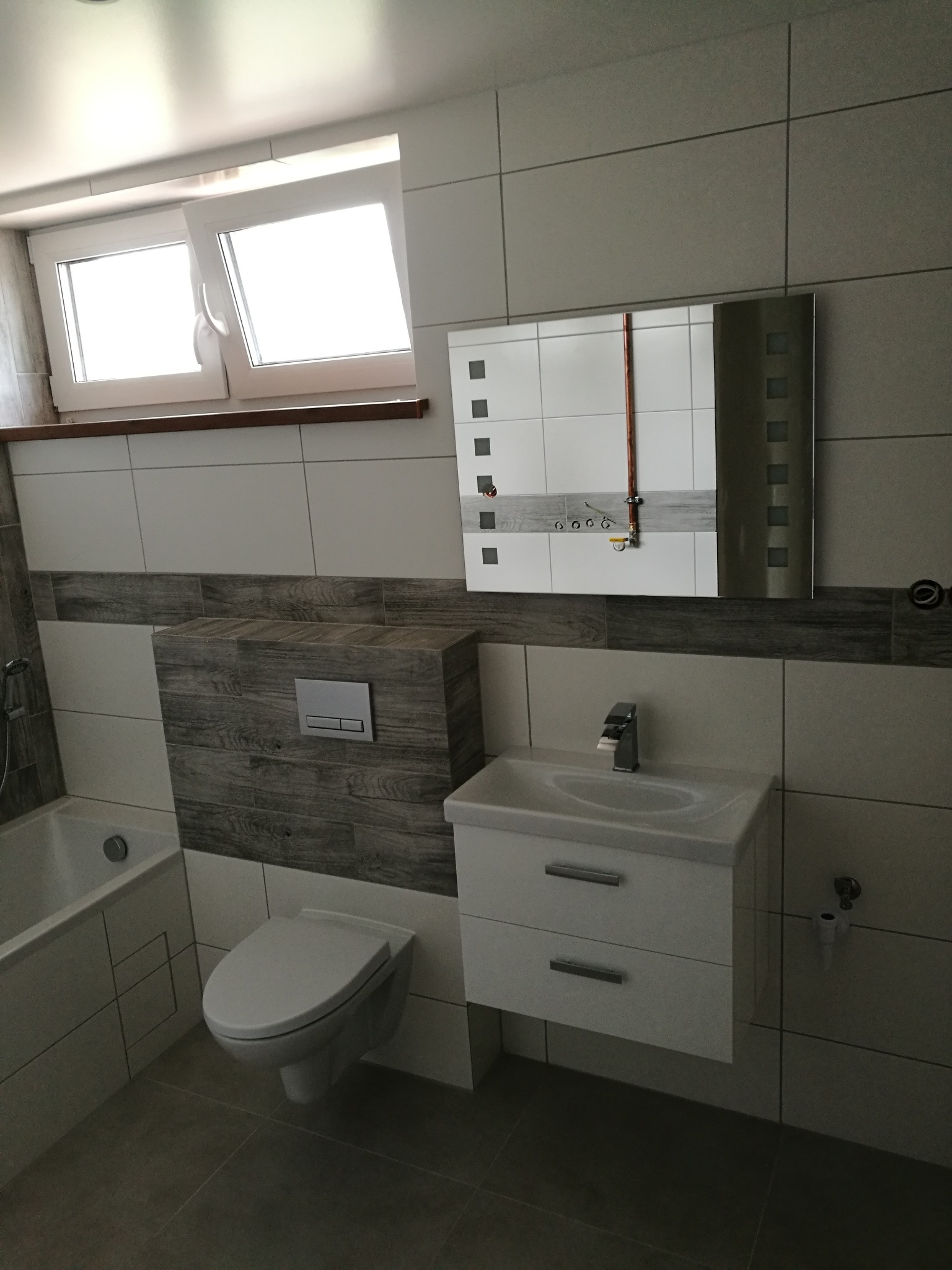 Wykończona łazienka z podwieszaną toaletą, białą umywalką z szafką i lustrem z elementami dekoracyjnymi, widoczne okno z drewnianym parapetem i szare płytki.