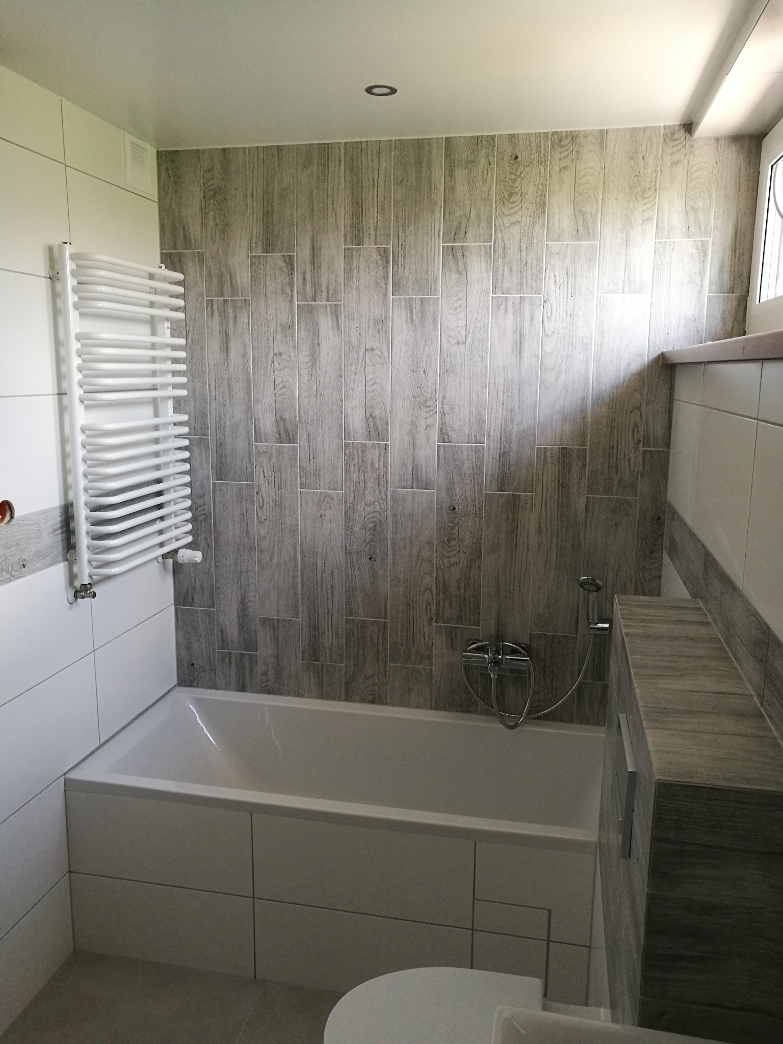 Biała wanna obudowana płytkami, grzejnik drabinkowy na ścianie z białych płytek, ściana wyłożona płytkami imitującymi drewno w odcieniach szarości, bateria prysznicowa.