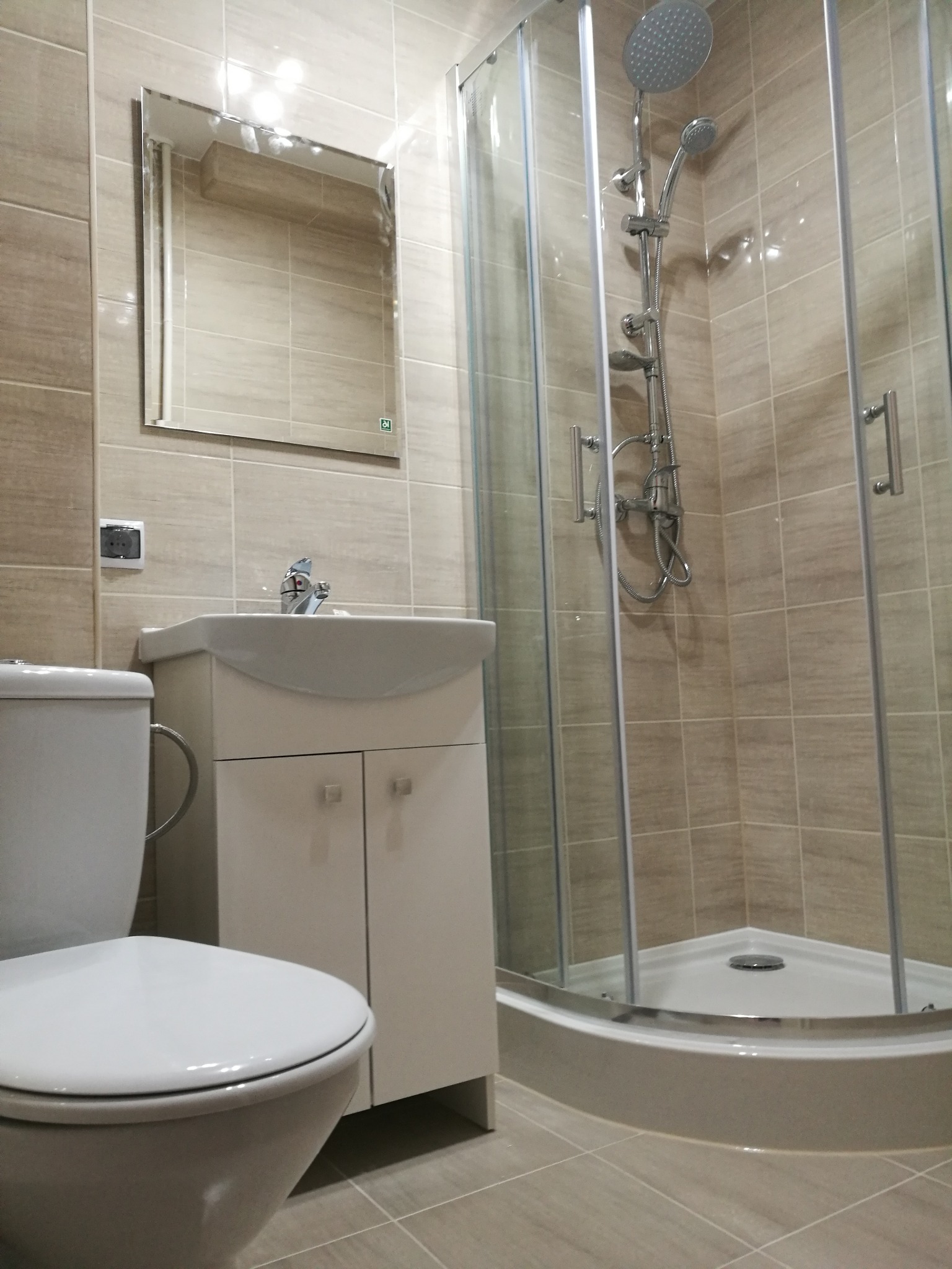 Jasna łazienka z kabiną prysznicową, umywalką z szafką i toaletą, wykończona płytkami w odcieniach beżu.