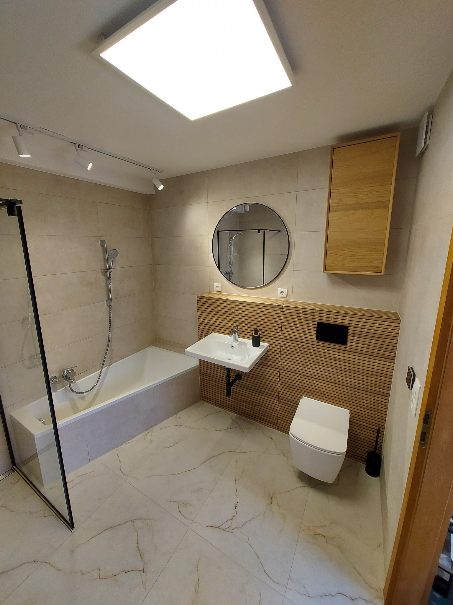 Jasna łazienka po remoncie: beżowe płytki imitujące marmur na podłodze i ścianach, wanna z czarną kabiną, umywalka z okrągłym lustrem i drewniane lamele na ścianie.