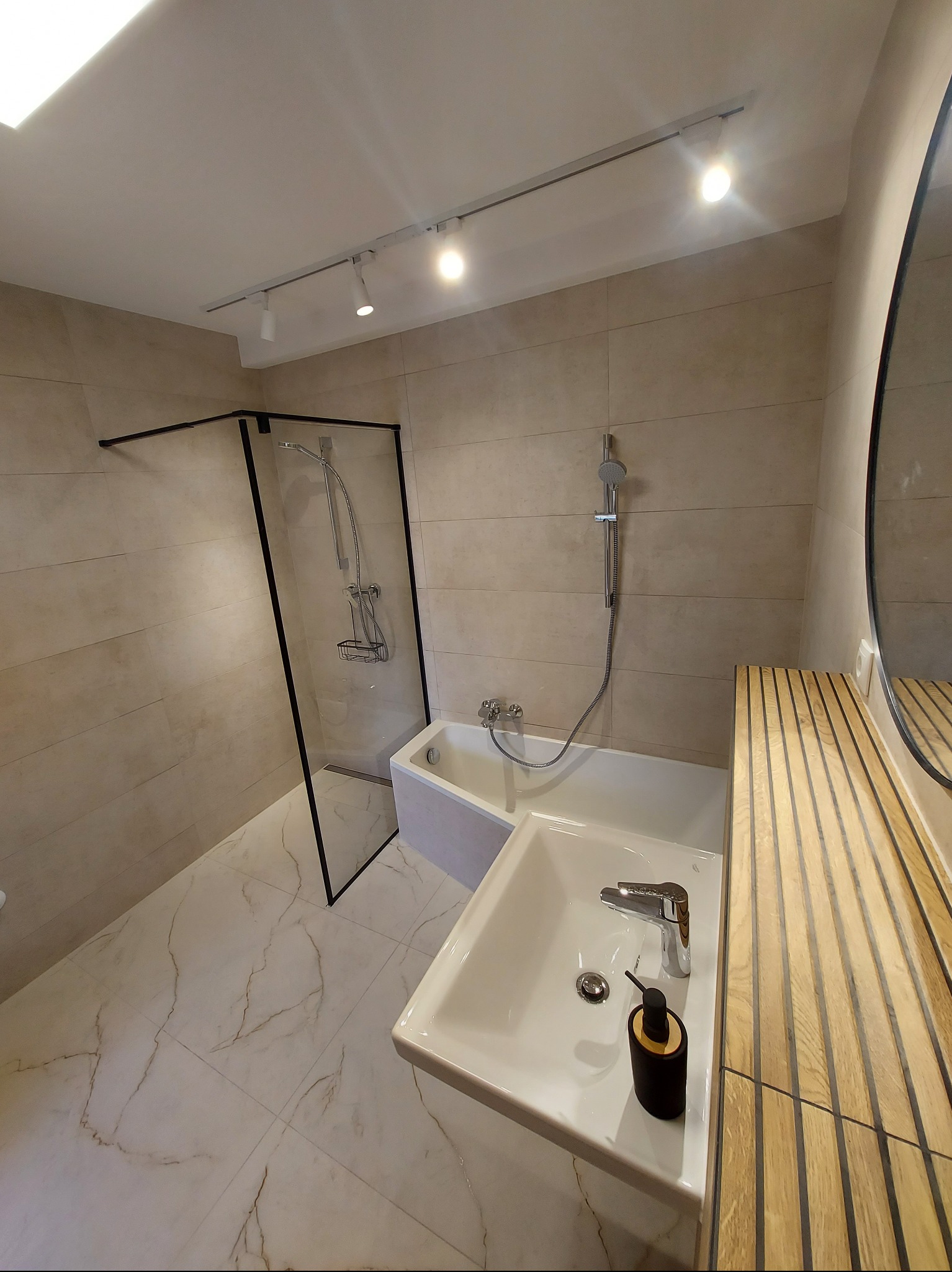 Nowoczesna łazienka po remoncie: kabina prysznicowa typu walk-in z czarną ramą, biała umywalka z drewnianym blatem, marmurowe płytki podłogowe i oświetlenie punktowe.