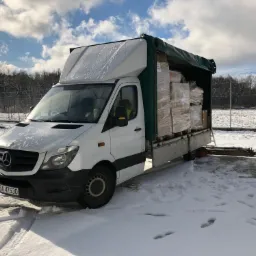 Biały bus marki Mercedes, częściowo załadowany paczkami owiniętymi folią stretch, stoi na śniegu w pobliżu ogrodzenia z drutu kolczastego w słoneczny dzień.