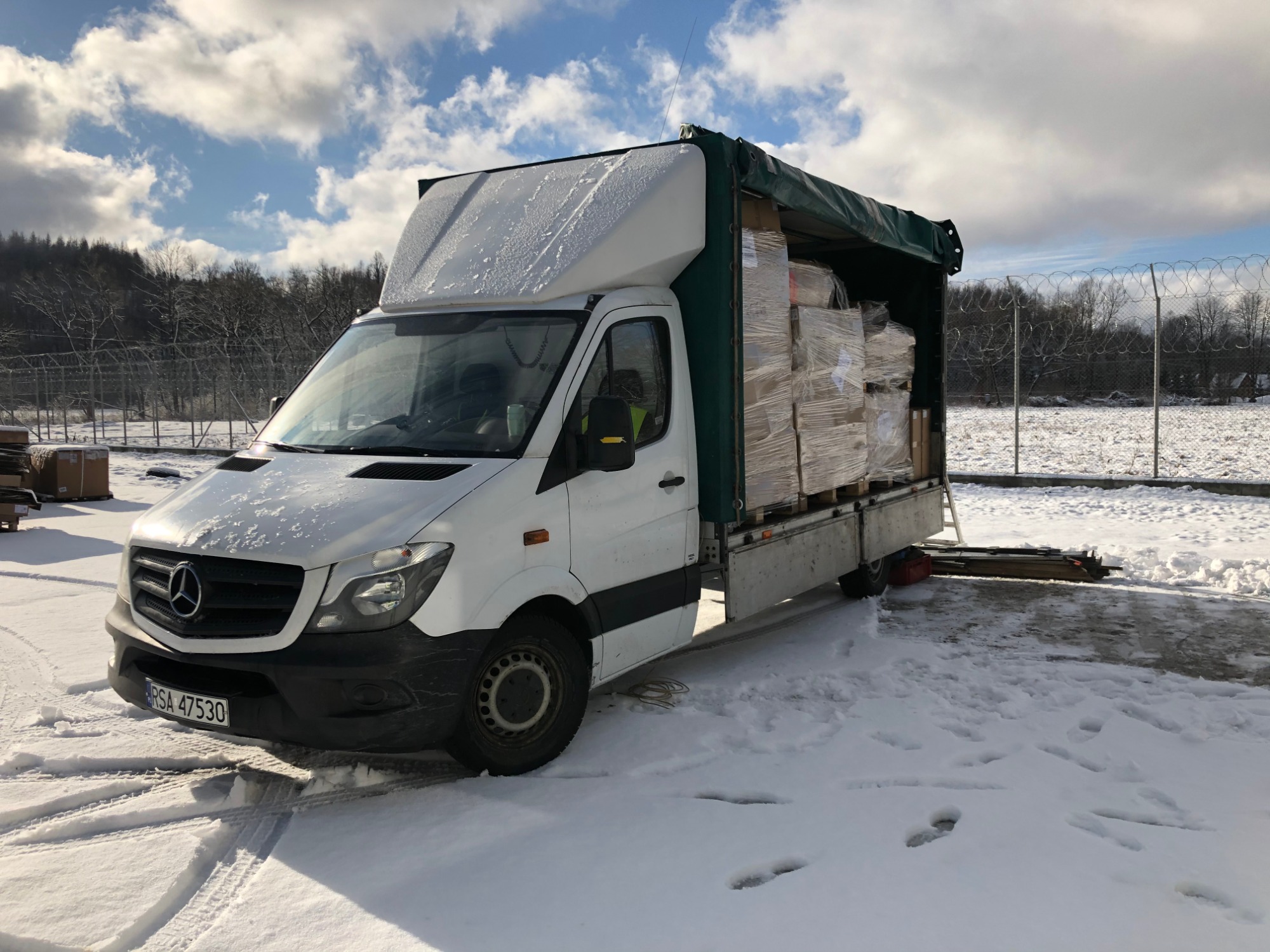 Biały bus marki Mercedes, częściowo załadowany paczkami owiniętymi folią stretch, stoi na śniegu w pobliżu ogrodzenia z drutu kolczastego w słoneczny dzień.