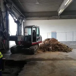 Mini koparka gąsienicowa wewnątrz budynku przemysłowego, obok wykopu i pryzmy ziemi, widoczne oświetlenie sufitowe i częściowo ogrodzenie.