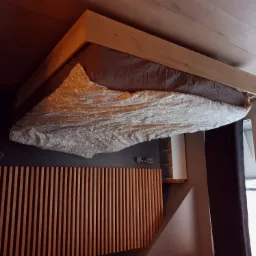 Drewniane łóżko z ramą z jasnego drewna, pościel w delikatny wzór, pionowe panele drewniane na ścianie za łóżkiem, widoczne gniazdka elektryczne.