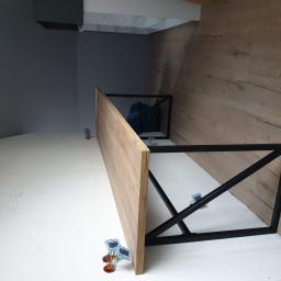 Forma drewna - Fragment blatu drewnianego z metalowym stelażem w trakcie montażu, widoczne elementy mocujące i ściana wykończona panelami drewnopodobnymi.