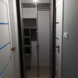 Wąska szafa wnękowa z otwartymi drzwiami w korytarzu, widoczne p&oacute;łki i drążek na ubrania, oświetlenie sufitowe.