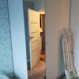 Forma drewna - Biała szafa z lustrzanymi drzwiami, w których odbija się otwarte drzwi do innego pomieszczenia. Obok szafy leżą elementy montażowe w folii i torbie.