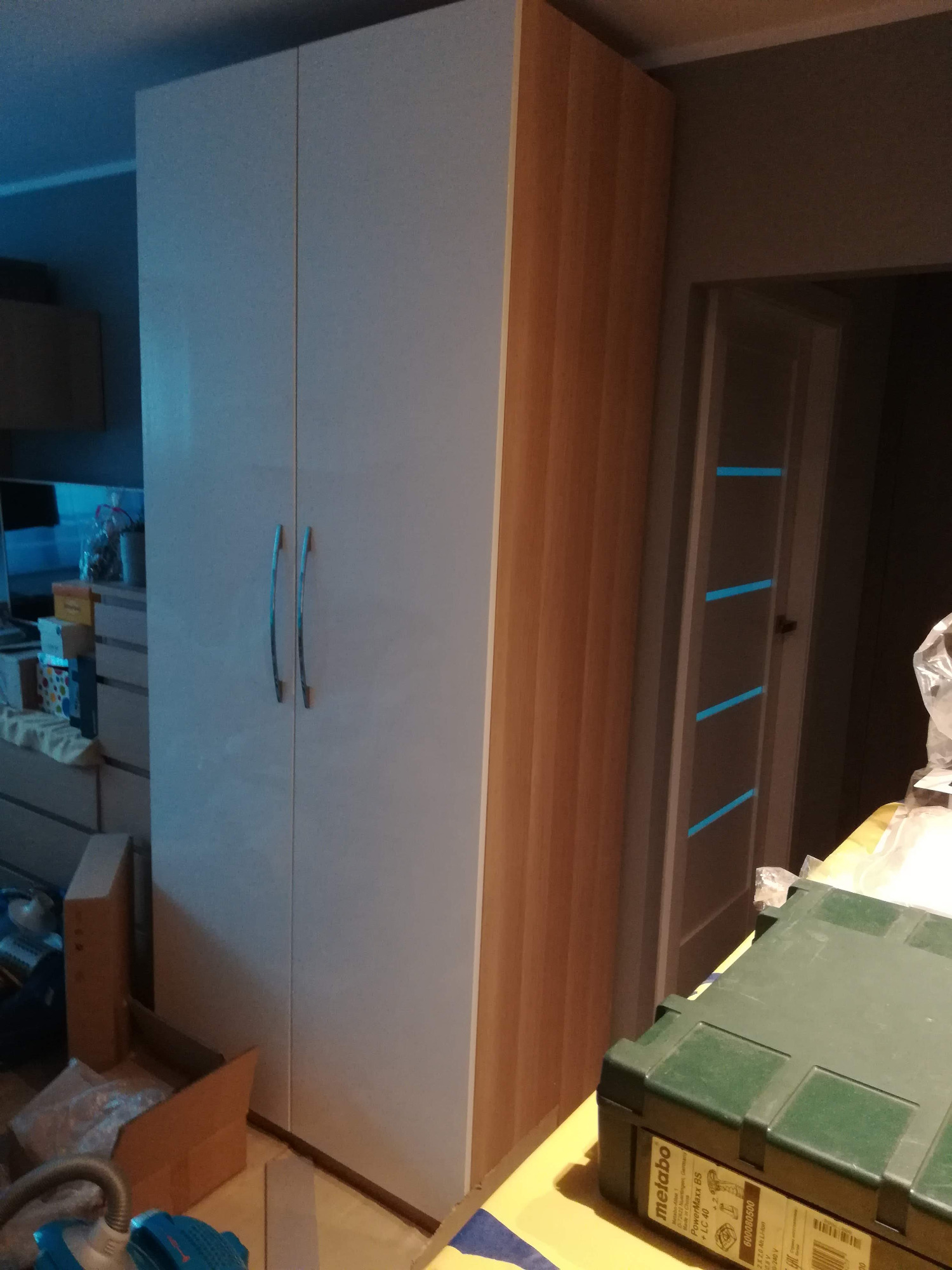 Dwudrzwiowa szafa z białymi frontami i bokami imitującymi drewno, widoczna w trakcie montażu, z narzędziami Metabo w zielonej skrzynce na pierwszym planie.