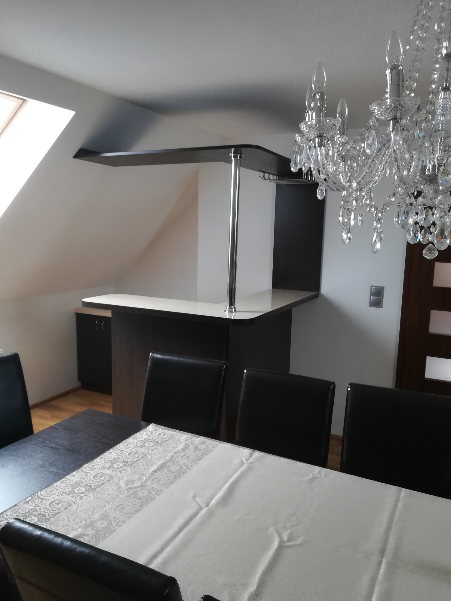 Kraków: Nowoczesny barek z blatem i metalową nogą, zamontowany w pomieszczeniu ze skosami, oświetlony kryształowym żyrandolem, widok zza stołu z krzesłami.