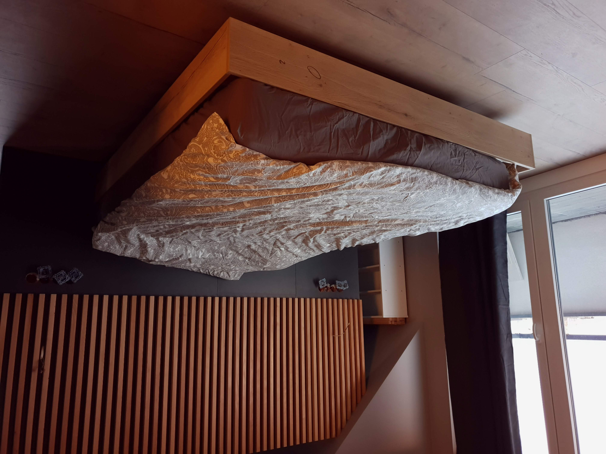 Drewniane łóżko z ramą z jasnego drewna, pościel w delikatny wzór, pionowe panele drewniane na ścianie za łóżkiem, widoczne gniazdka elektryczne.