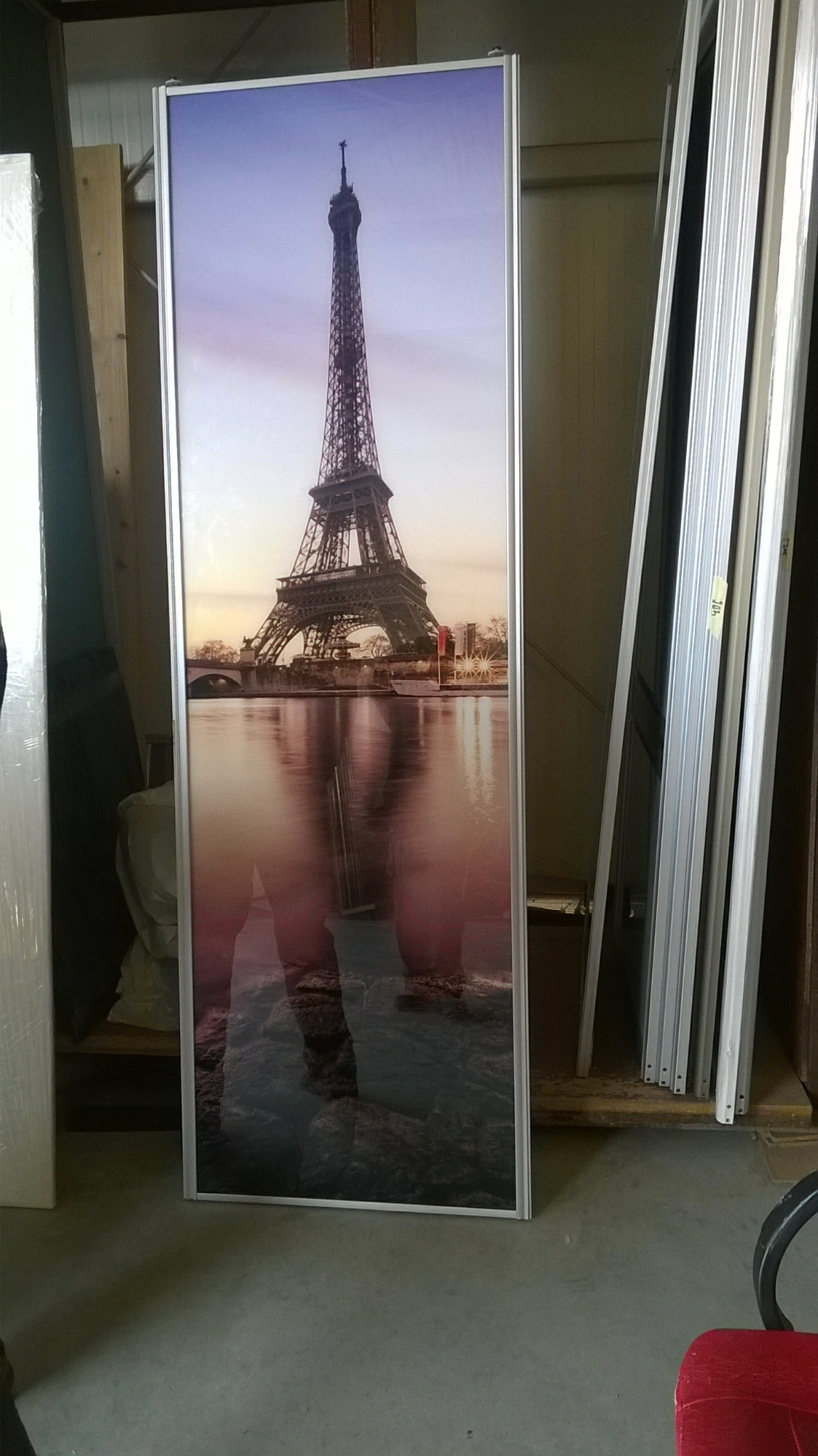 Wysoki, wąski panel z nadrukiem wieży Eiffla w srebrnej ramie, oparty o ścianę w warsztacie, obok stosu aluminiowych profili.