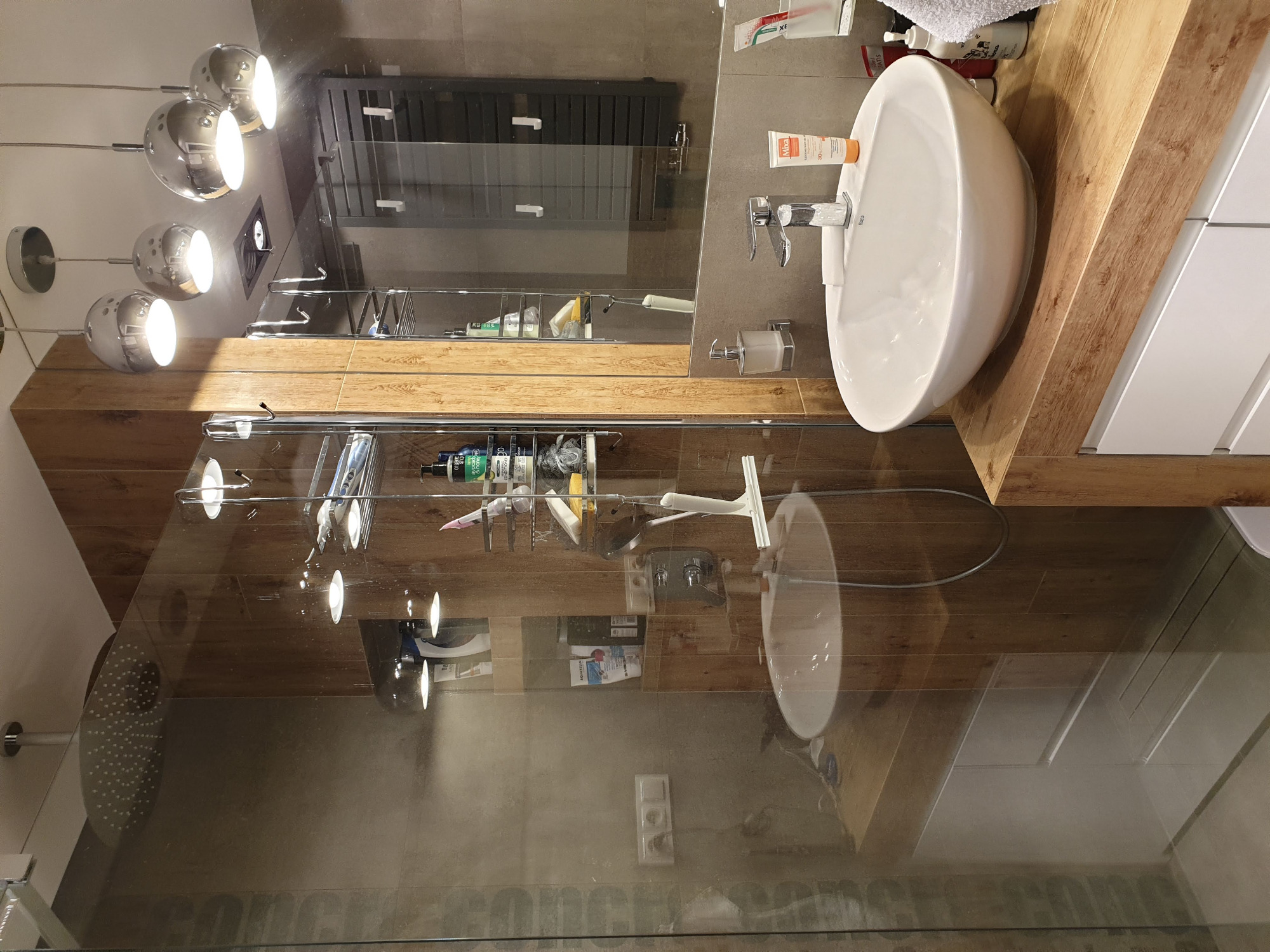 Nowoczesna łazienka z drewnianym blatem, białą umywalką i szklaną kabiną prysznicową, oświetlona kulistymi lampami.