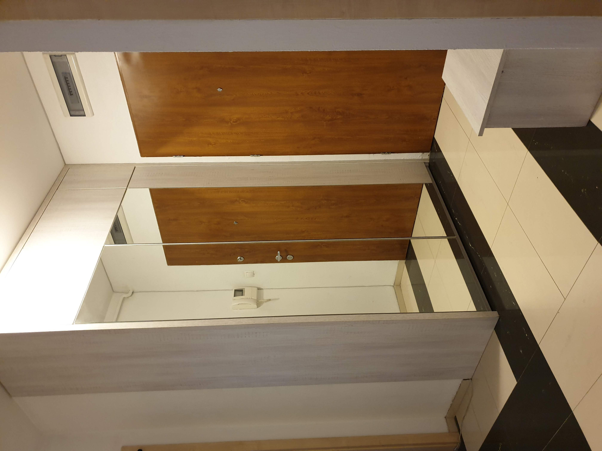 Wnętrze przedpokoju z zabudową szafy z lustrzanymi drzwiami przesuwnymi i drewnianą okleiną, widoczny fragment podłogi w czarno-białe pasy.