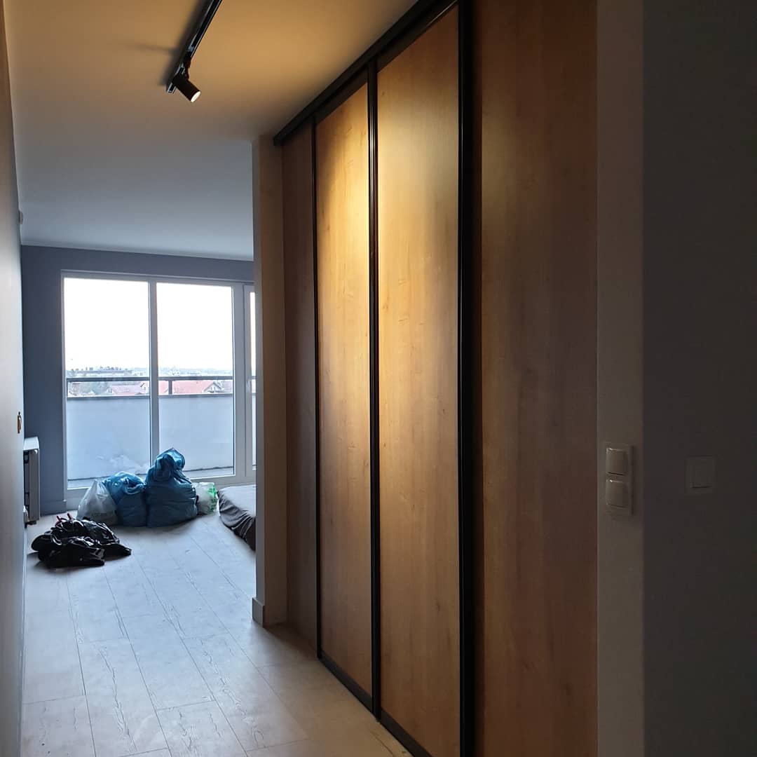 Wysoka szafa przesuwna z drewnianymi frontami i czarnymi obramowaniami, widok z korytarza, w tle okno balkonowe i worki z odpadami.