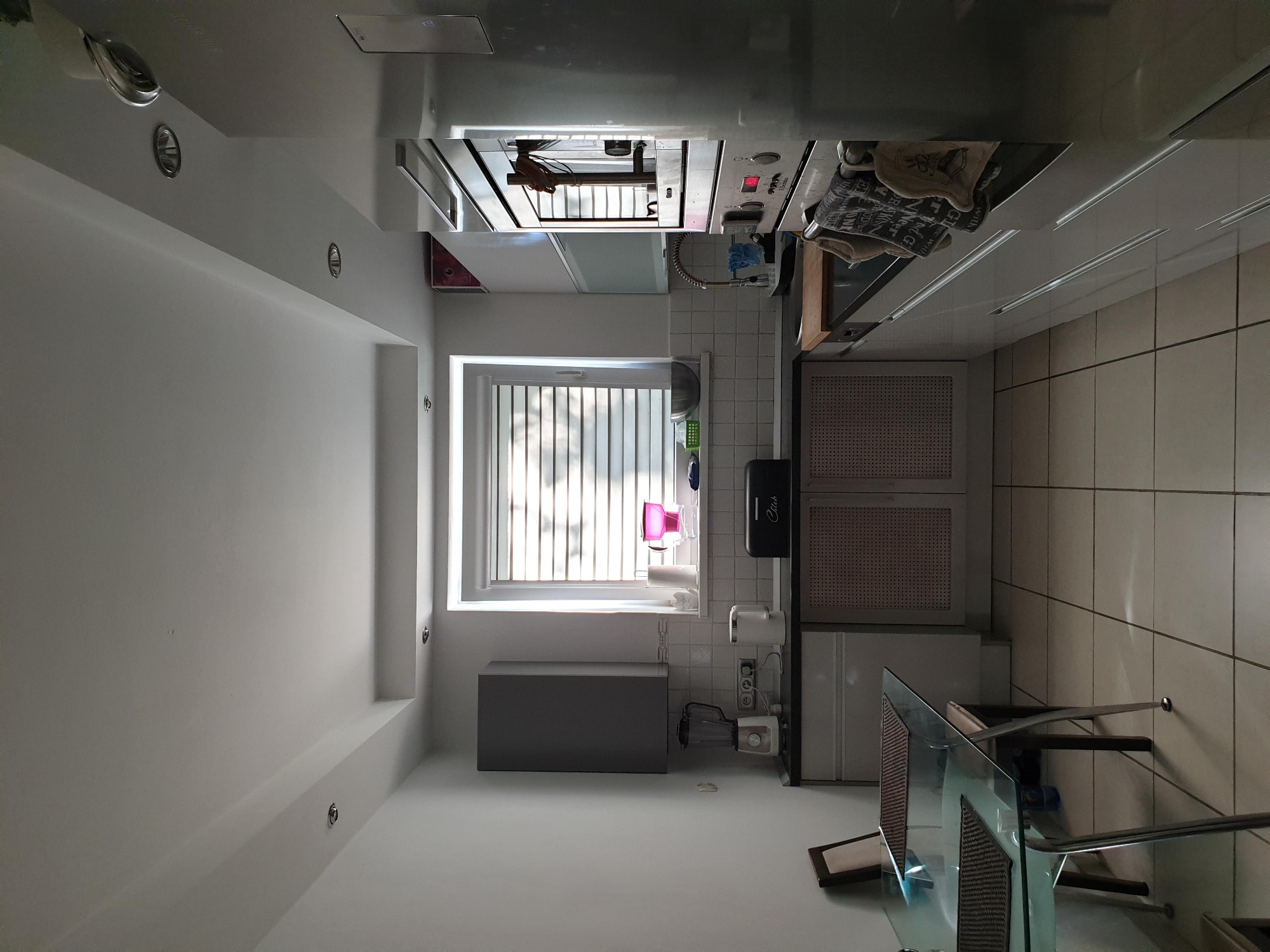 Jasna kuchnia z białymi ścianami i oknem z żaluzjami, widoczny fragment blatu i szafek kuchennych, minimalistyczny design.