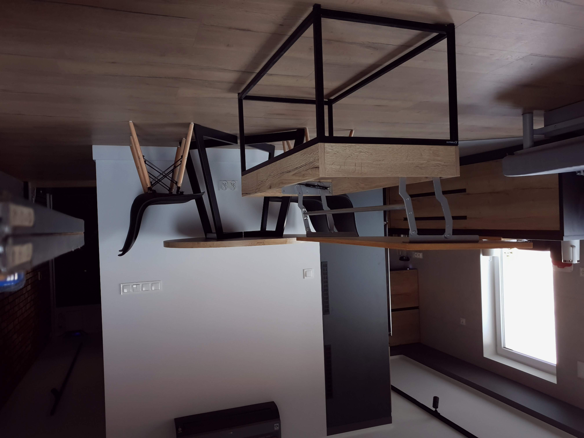 Nowoczesna jadalnia z drewnianym stołem na metalowym stelażu i krzesłami z czarnymi siedziskami, widok na białą ścianę z włącznikami i okno.