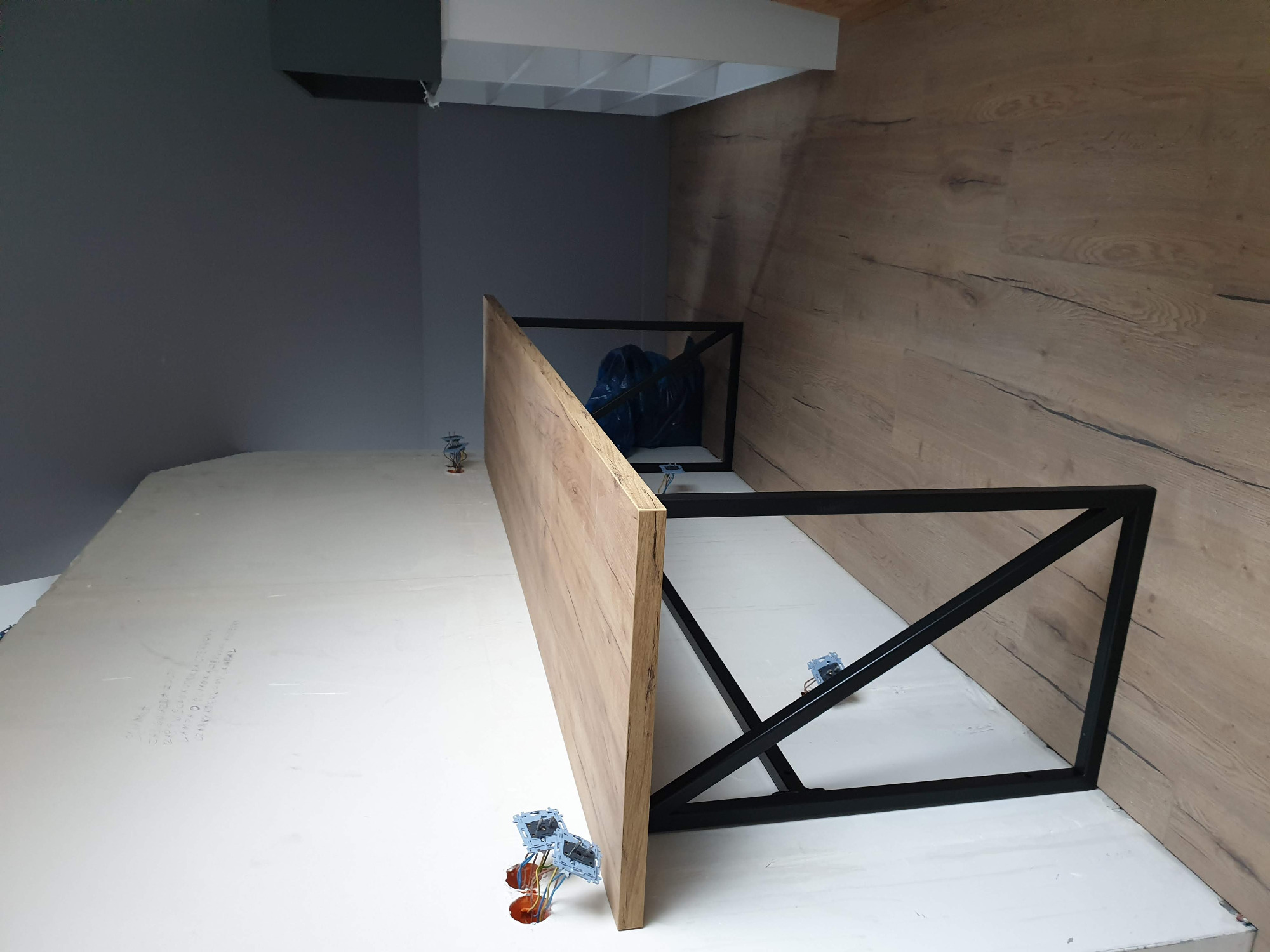 Fragment blatu drewnianego z metalowym stelażem w trakcie montażu, widoczne elementy mocujące i ściana wykończona panelami drewnopodobnymi.