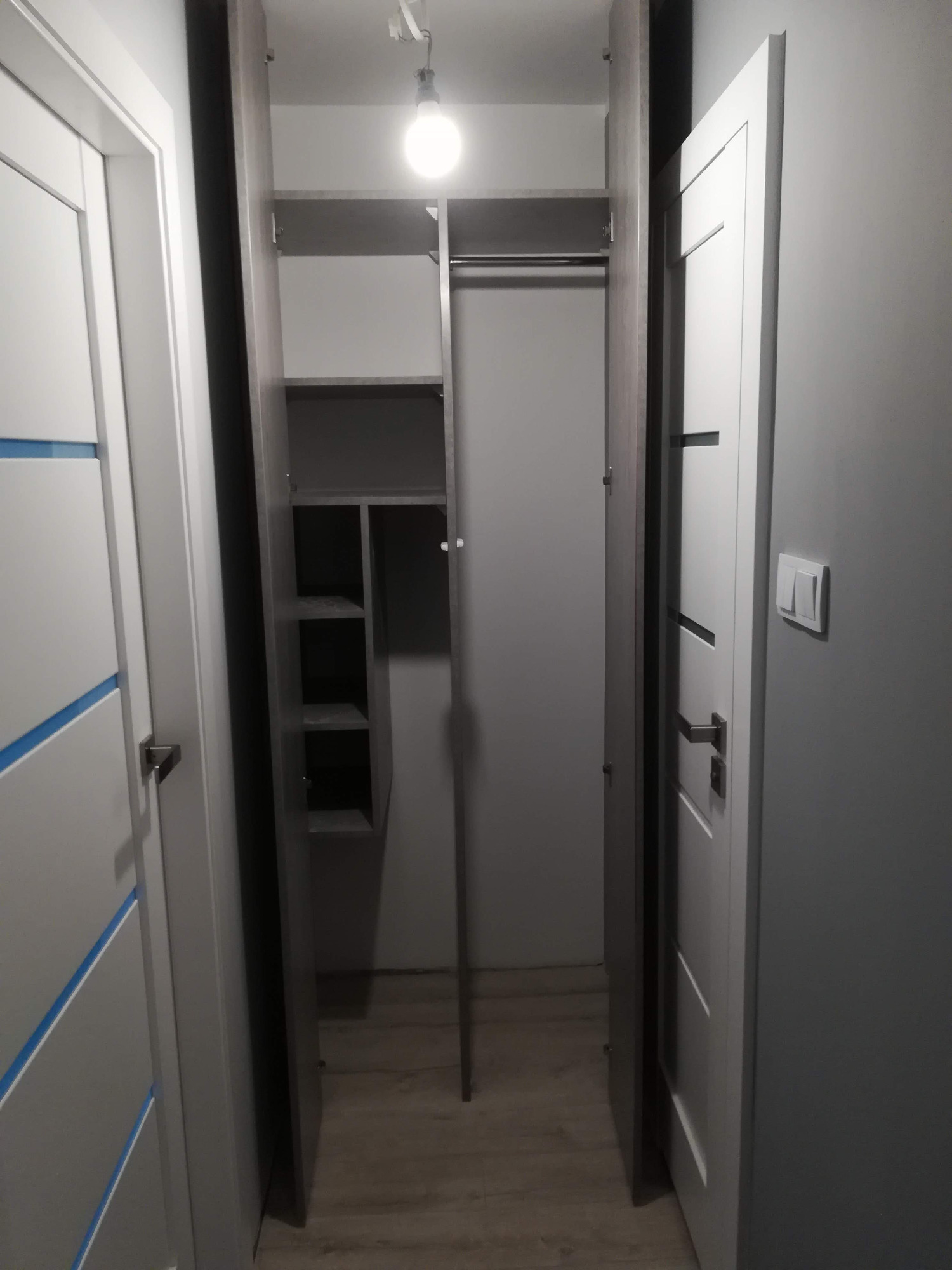 Wąska szafa wnękowa z otwartymi drzwiami w korytarzu, widoczne półki i drążek na ubrania, oświetlenie sufitowe.