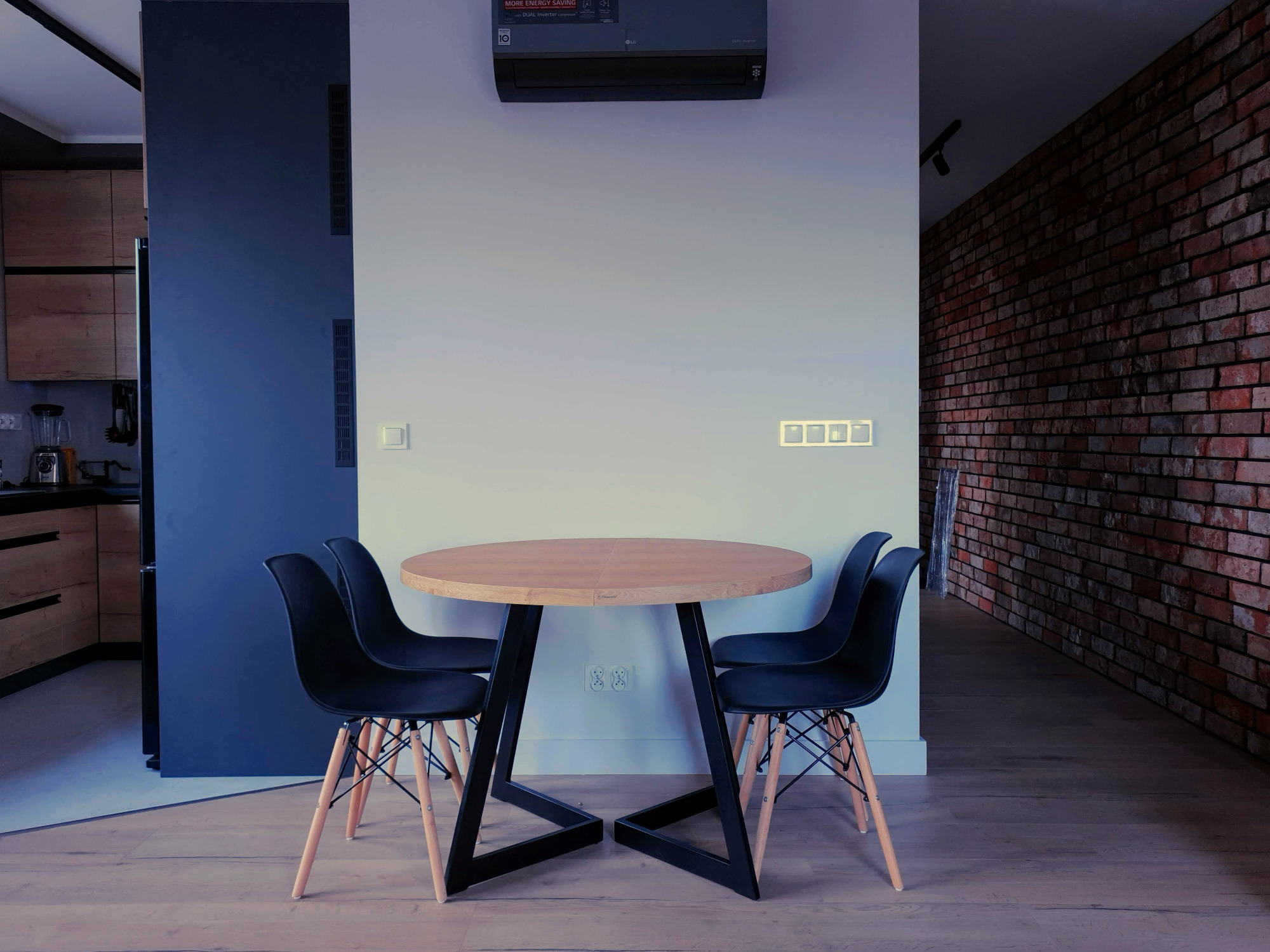 Okrągły stół z jasnego drewna z czarną, geometryczną podstawą i czterema czarnymi krzesłami na drewnianej podłodze, na tle ściany z cegły i kuchni w tle.