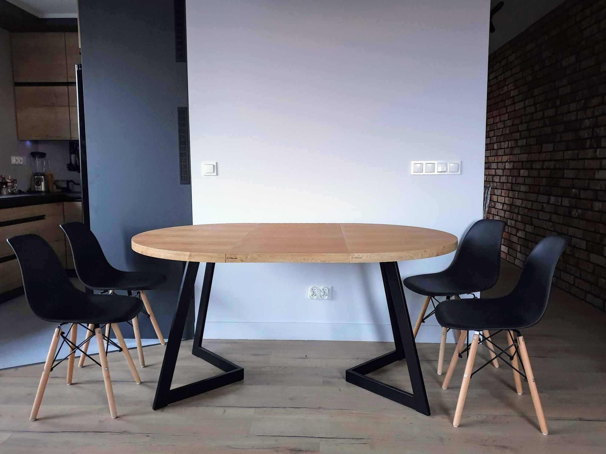 Owalny rozkładany stół z jasnego drewna na czarnych, geometrycznych nogach, otoczony czarnymi krzesłami z drewnianymi nogami, na tle białej ściany i fragmentu ściany z cegły.