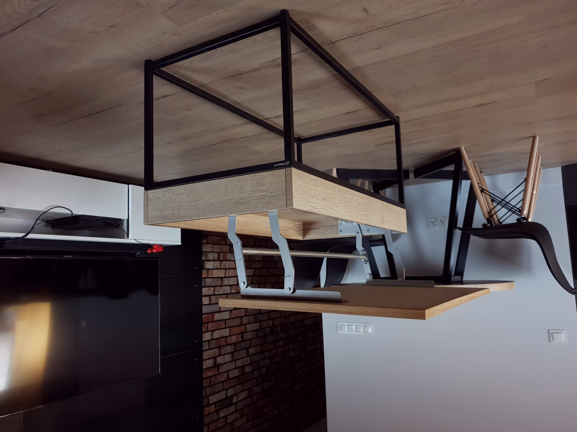 Rozkładany blat biurka zamontowany pod szafką, z metalową konstrukcją nośną, widok na ceglaną ścianę i czarny telewizor w tle.