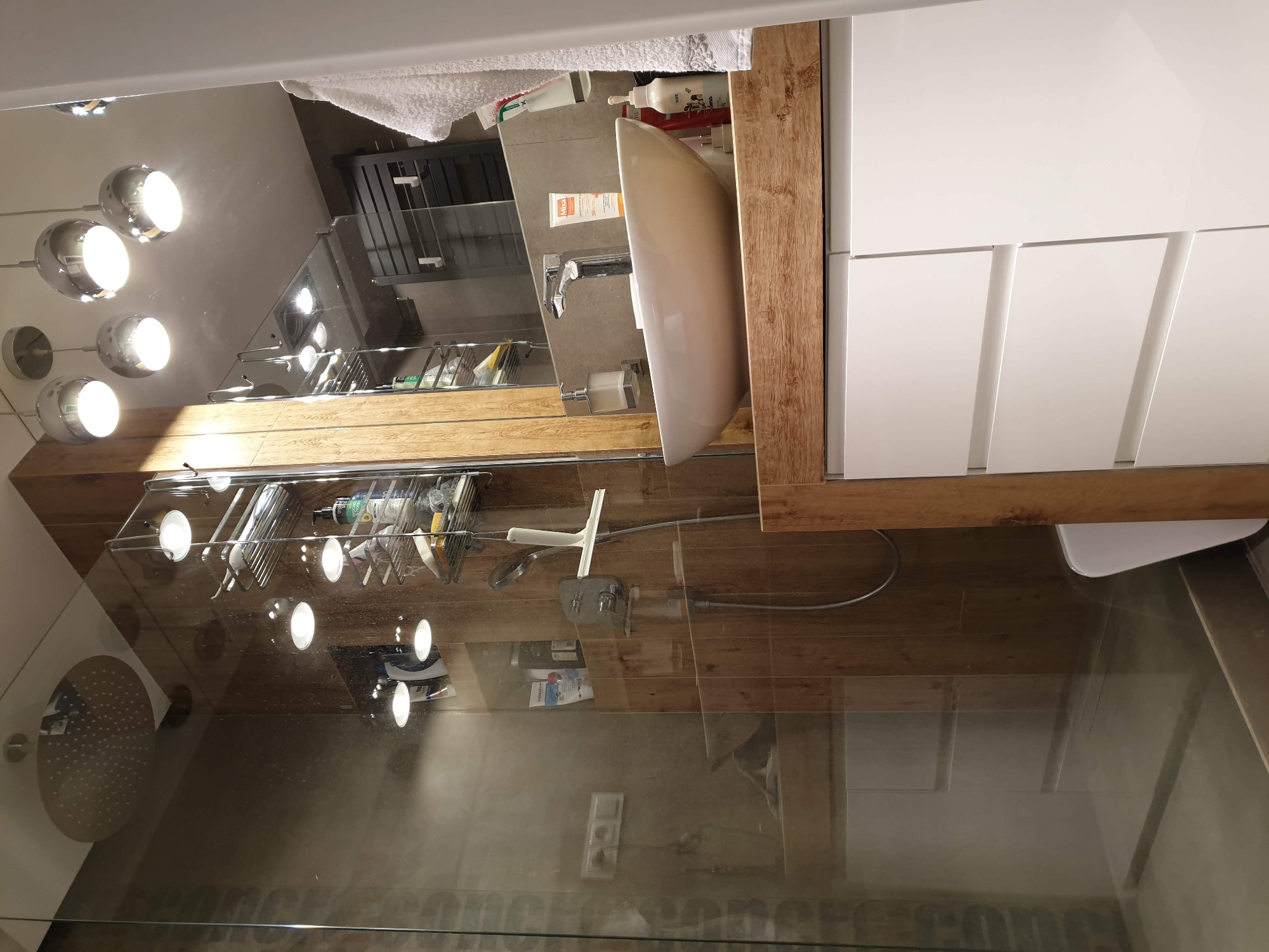 Nowoczesna łazienka z białymi szafkami pod umywalką, drewnianym blatem i szklaną kabiną prysznicową z chromowaną deszczownicą