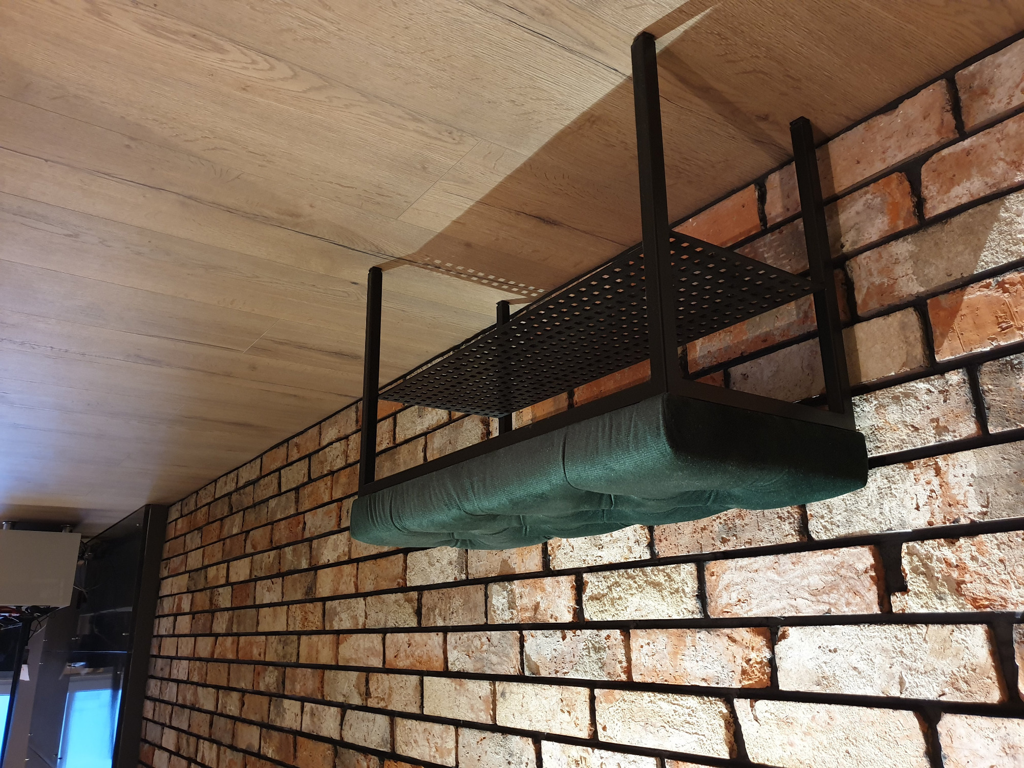 Loftowy wieszak ścienny z tapicerowanym siedziskiem w kolorze butelkowej zieleni, zawieszony na tle ceglanej ściany i drewnianego sufitu.