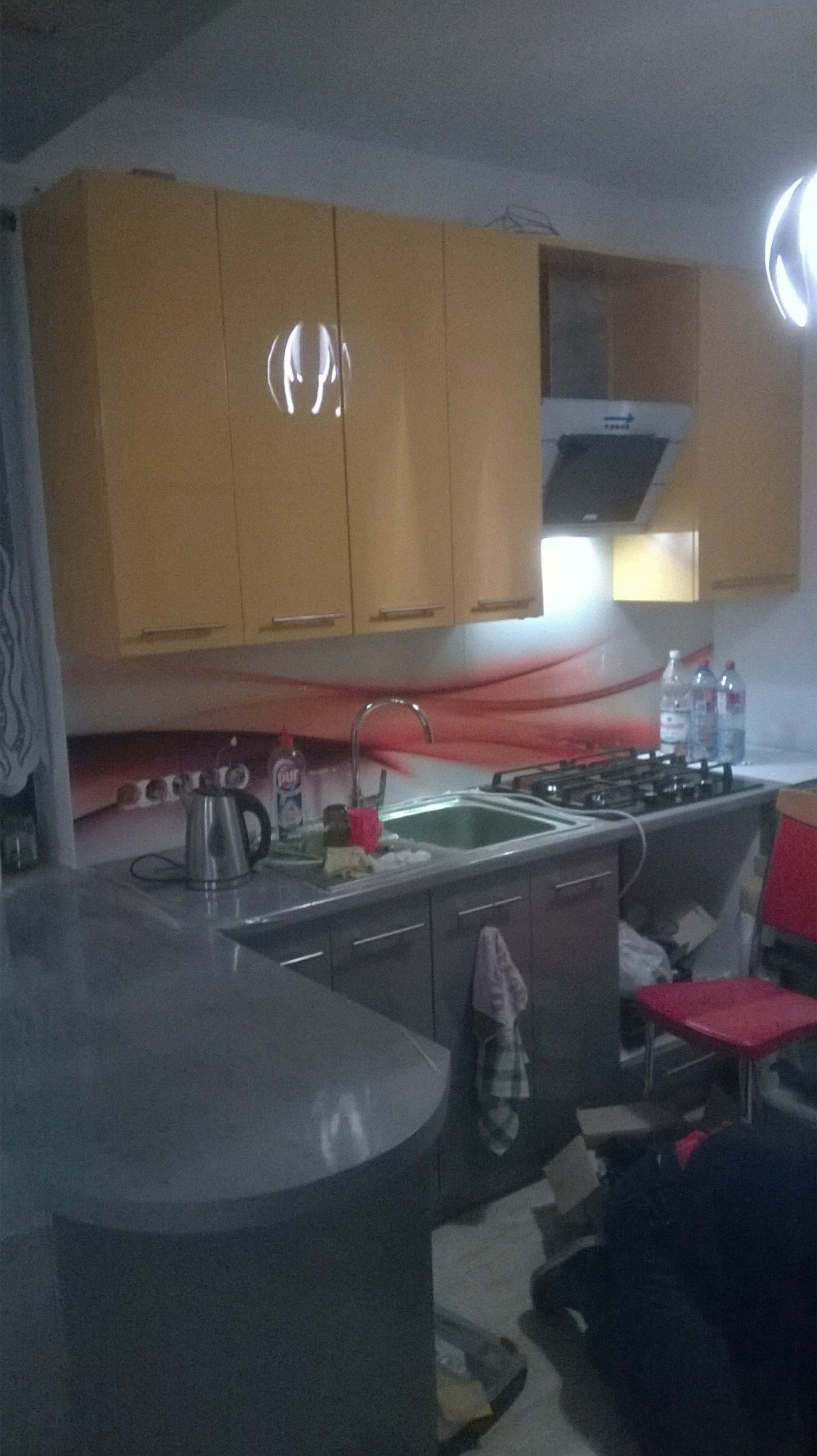 Żółte szafki kuchenne z błyszczącymi frontami, srebrny zlew i płyta gazowa na tle czerwonego panelu ściennego, widoczny fragment blatu barowego i czerwonego krzesła.